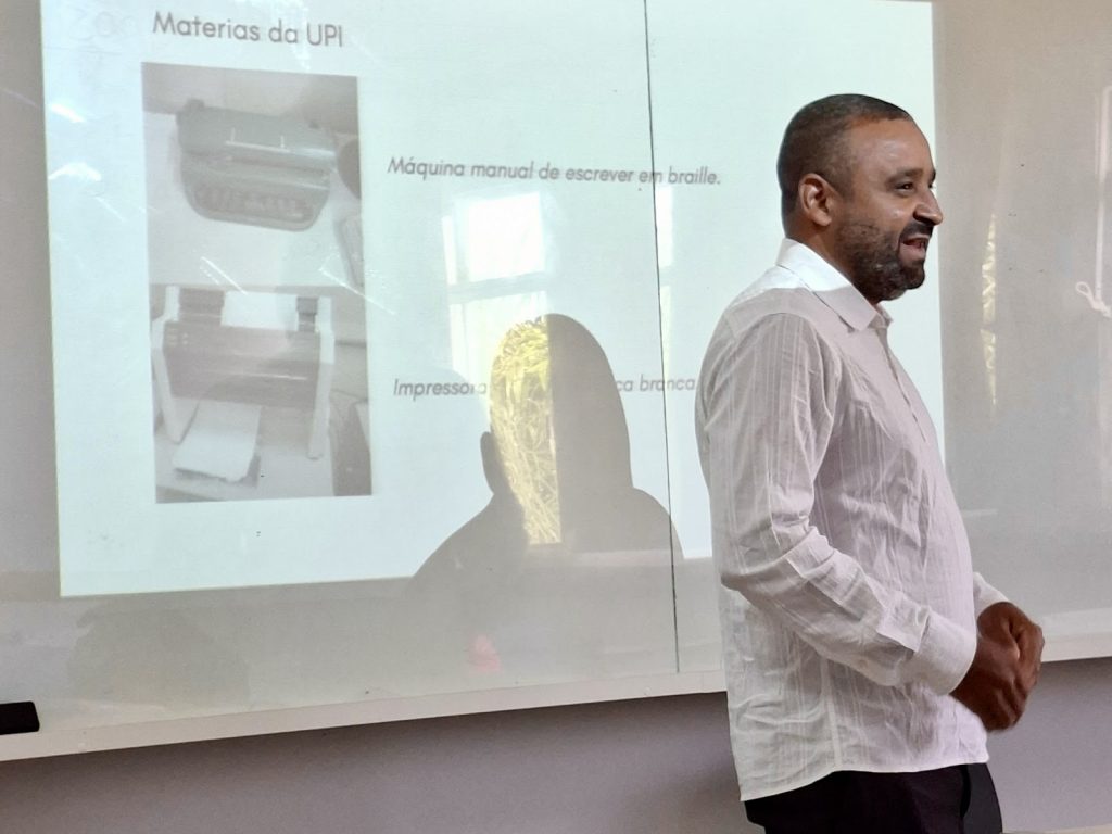 Edmilson é um jovem negro, cabelos rentes à cabeça, barba e bigode curtos. Está costas para um telão.